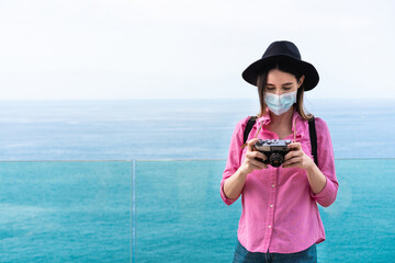 Young tourist woman using vintage old camera while wearing face mask with ocean view in background - Travel and safety measures during coronavirus outbreak concept - Focus on face