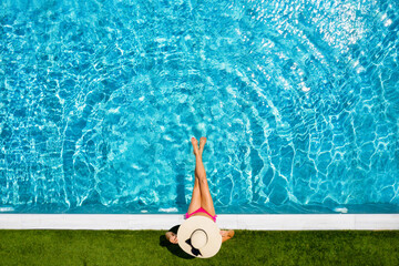 Top Ansicht einer Frau mit Hut die am Rande eines Swimming Pools sitzt und das blaue, kühle Wasser...