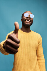 Naklejka premium portrait photo of a dark-skinned handsome guy with a beard on a blue background in a yellow sweater, transparent glasses, he looks at the camera, smiles and keeps his thumb up