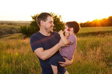 Dad and son walk in a yellow summer field in the evening at sunset and eat candy on a stick. Father's Day, love in the family, the role of the father in raising the child.