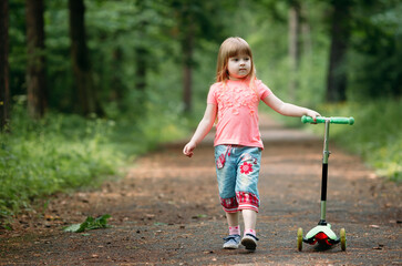 Happy little girl with long hair stands and holds a scooter in a summer park, looks to the side. Copy space