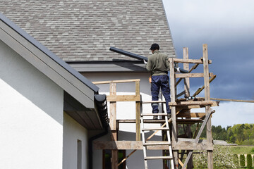 Construction worker installing plastic roof rain gutter gray shingles roof with wooden eaves while standing on scaffolding, back view. Home improvement and diy concept.
