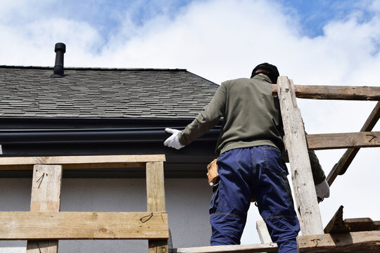 Construction worker installing plastic roof rain gutter gray shingles roof with wooden eaves while standing on scaffolding, back view. Home improvement and diy concept.
