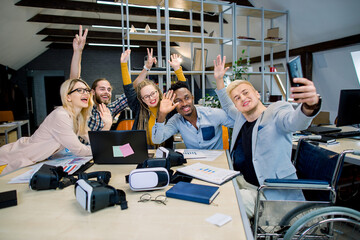 Handsome joyful man in wheelchair sitting at desk with his colleagues, and making funny selfie photo on smartphone. Group of multiethnic hipster coworkers posing for photo and waving