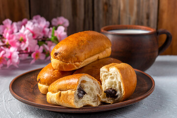 Breakfast, delicious rolls with chocolate filling on a plate, milk in a clay mug on a light textured table, wooden background decorated with a sprig of cherry blossoms