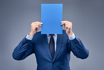 A businessman in a blue suit on a gray-blue background holds a sheet of clean blue paper at head level. Concept of product or service presentation, sales, or performance. Free space for text.
