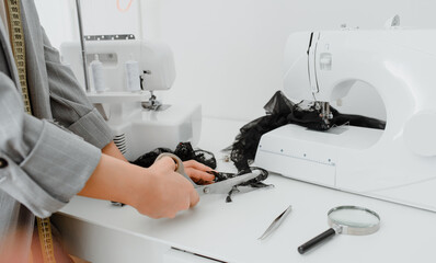 Young woman tailor is cutting with scissors a piece of black material in her white mini workshop.