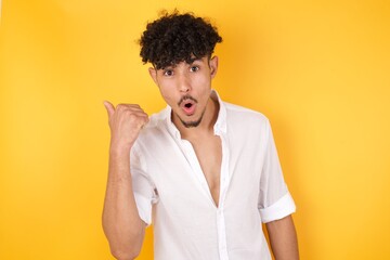 Horizontal shot of pretty young joyful shocked male points with thumb away, indicates something on blank wall isolated over gray background. Check this out. Ethnicity concept.