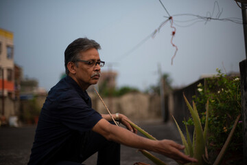 An old/aged Indian Bengali man in blue shirt is gardening on a rooftop under the open sky. Indian lifestyle and seniors