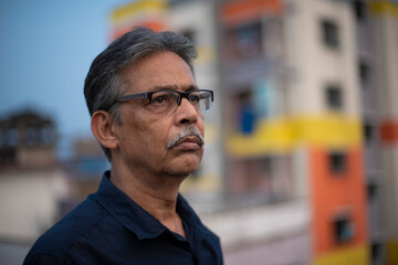 Close up portrait of an An old/aged Indian Bengali man in blue shirt is standing on a rooftop under the open sky in front of a urban landscape. Indian lifestyle and seniors