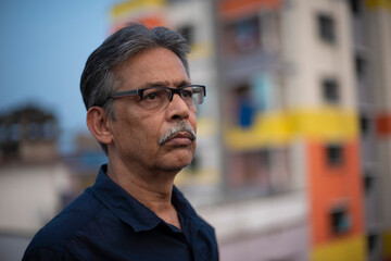 Close up portrait of an An old/aged Indian Bengali man in blue shirt is standing on a rooftop under the open sky in front of a urban landscape. Indian lifestyle and seniors