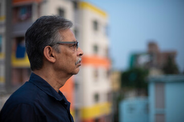 Close up portrait of an An old/aged Indian Bengali man in blue shirt is standing on a rooftop under the open sky in front of a urban landscape. Indian lifestyle and seniors