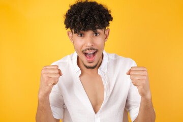 Portrait of Handsome caucasian blond male looks with excitement at camera, keeps hands raised over head, notices something unexpected, isolated over gray wall. Lovely man reacts on sudden news.