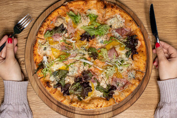 Big delicious pizza and women's hands with a fork and a knife on a wooden background. National Italian cuisine, restaurant