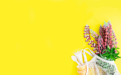 A string bag with a bouquet of fresh pink lupins, cardboard disposable cups and wooden spoons, forks, knives on a yellow background. The concept of zero waste and no plastic with space for your text