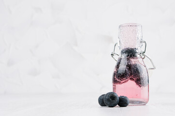 Refreshing cold fruit pink drink with blueberry, bright bubbles in transparent yoke bottle in elegant white interior on wood table, copy space.