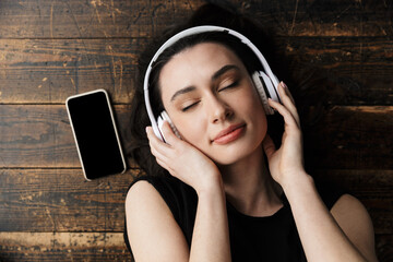 Photo of young woman wearing headphones lying on floor with cellphone