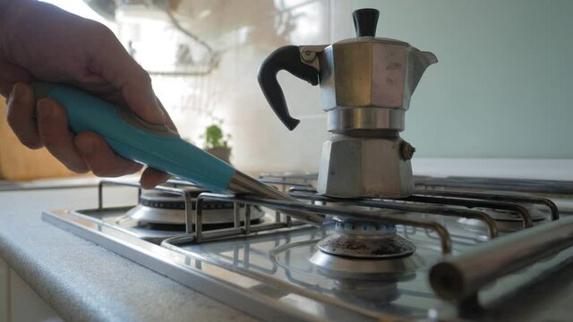 Man puts the moka pot, the traditional Italian machine for making coffee, on the cooker of a kitchen illuminated by the light coming from a window, then lights the flame with a lighter