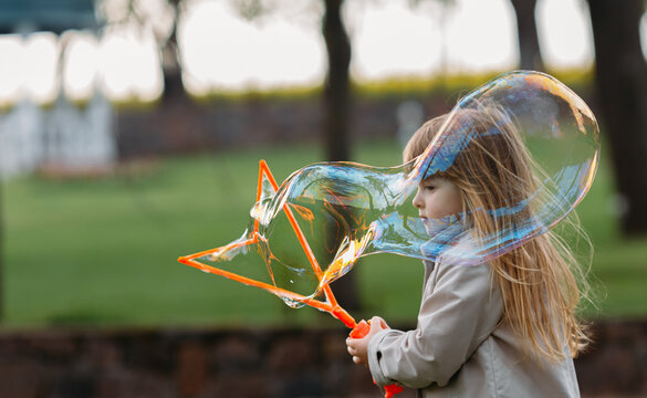 Portrait of a little cute blonde girl having fun and joy blowing big soap bubbles playing on the street. Cheerful charming child enjoys a healthy summer game