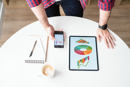 An anonymous man is holding a mobile phone. The man is working from home. The tablet and the phone show graphics. There is a mug of coffee in the desk