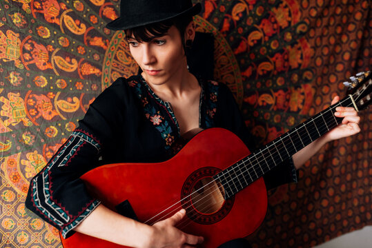 Delighted female hipster in trendy hat and dress leaning on colorful wall cover and playing acoustic guitar