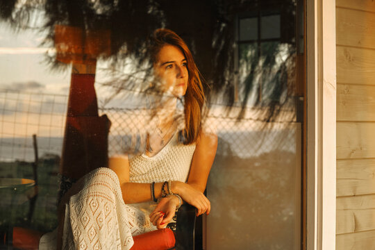 Serene female in knitted summer dress sitting in cozy armchair near window and admiring picturesque sunset while enjoying vacation in wooden cabin