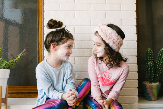 Joyful laughing girls in similar clothes and applied face mask sitting on wooden table at home having fun against brick white wall