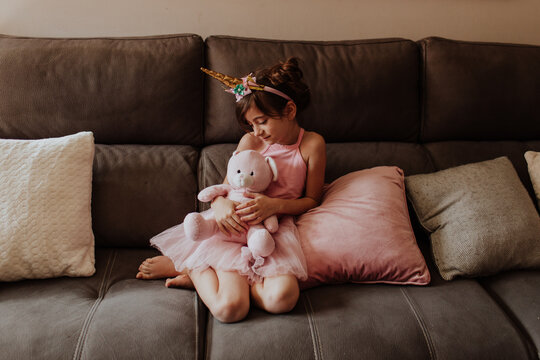 Barefoot girl in unicorn costume embracing plush toy while resting on comfortable couch at home