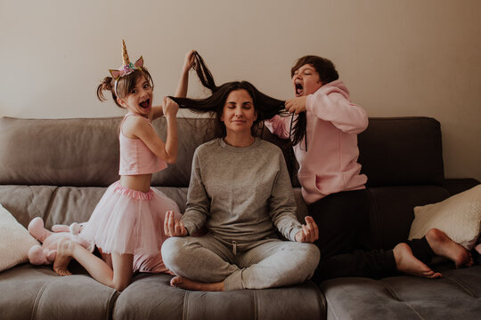 Excited little girl and teen boy pulling hair of adult woman sitting in Lotus pose on bed and meditating