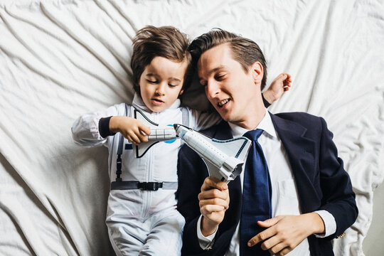 From above of cheerful boy in astronaut costume and dad in formal suit relaxing together in bedroom and playing with toy rockets