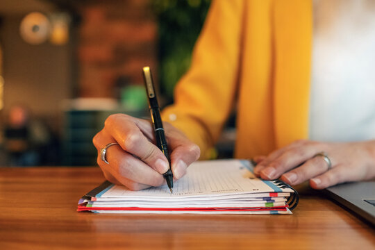 Crop unrecognizable businesswoman in elegant colorful clothes writing with pen in notebook while sitting at wooden table and using laptop in light contemporary office