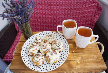 Two cups of black tea stand on a wooden tray, a bouquet of dried lavender. Fresh and fragrant cinnamon rolls close up lie on a plate with polka dots, beautiful morning. Close-up