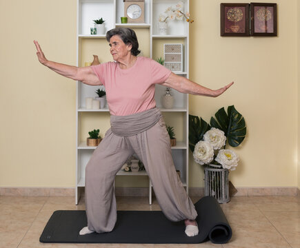 Full body of elderly lady in active wear practicing yoga pose while standing on mat near shelves with potted plants