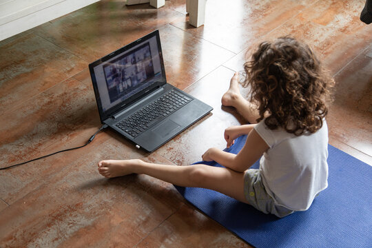 Back view of unrecognizable little girl watching online video tutorial on laptop while sitting on mat and learning yoga pose at home