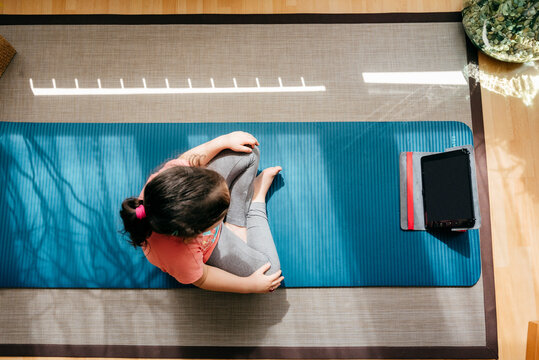 Top view of unrecognizable little girl sitting in lotus pose and meditating on mat at home while practicing yoga and watching video tutorials on tablet