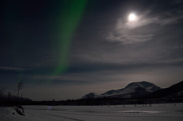 aurora borealis, northern light over snowy winter landscape with mountain and full moon at night