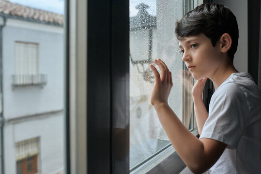 Bored child in casual clothing standing in living room and leaning on window while observing rain on street