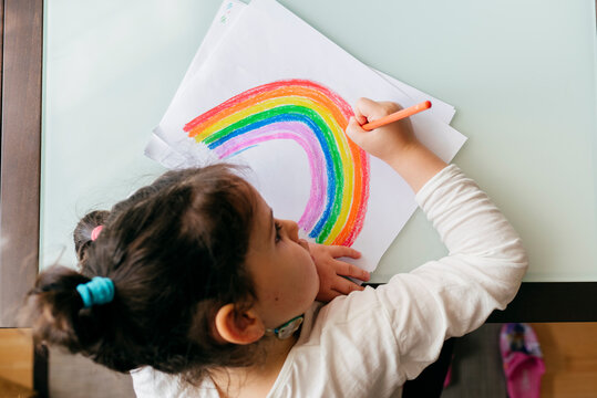 From above girl in casual clothes sitting on chair at wooden table and drawing colorful rainbow with pencil on paper