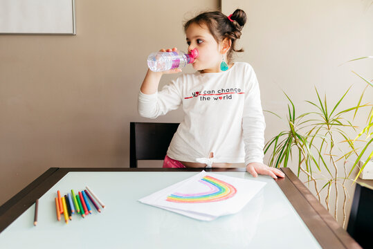 Positive girl in casual clothes drinking water from plastic bottle while standing near wooden table and drawing picture with colored pencils at home