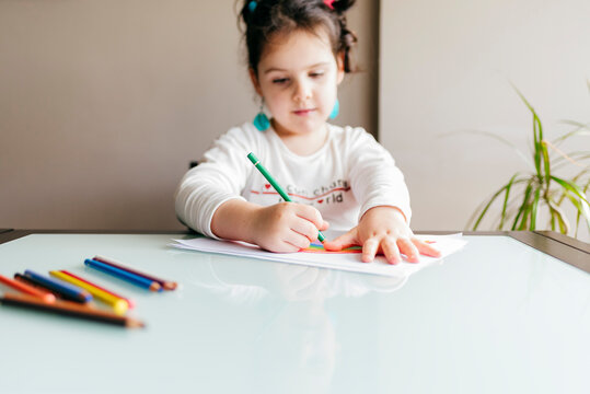 Girl in casual clothes sitting on chair at wooden table and drawing colorful rainbow with pencil on paper