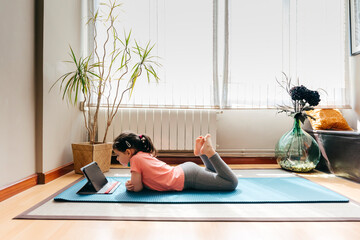 Side view of positive little girl lying down on mat near window at home resting after practicing yoga throughout video tutorials on tablet
