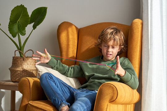 Cheerful preteen boy in casual clothes sitting in cozy armchair and playing interesting cats cradle game while resting at home