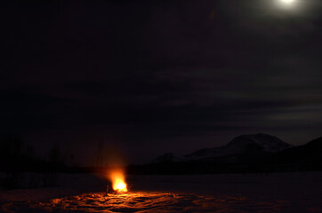 Fototapeta na wymiar burning fire on frozen river bed with majestic snowy mountain and full moon in the background