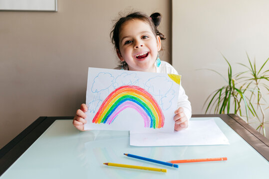 Smiling charming girl holding sheet of paper with colorful drawing while sitting at wooden table and smiling and looking at camera