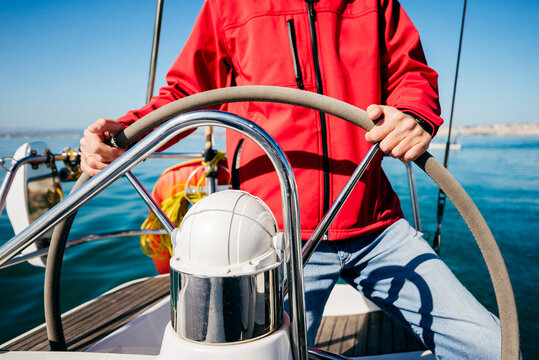 Crop unrecognizable man in red jacket and jeans standing at steering wheel while sailing in sea on modern vessel