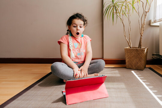 Surprised little girl sitting on mat near window at home resting after practicing yoga throughout video tutorials on tablet
