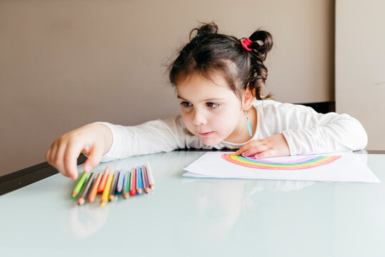 Concentrated little girl in casual clothes sitting at wooden table and taking colored pencil while drawing rainbow on paper at home