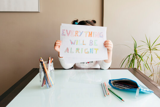 Anonymous girl sitting at wooden table with colored pencils and demonstrating drawing saying Everything will be alright at camera