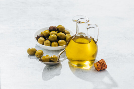 From above of glass jug with oil and small bowl and spoon with olives placed on white table