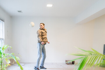 Skilled young man jumping performing mid-air trick with soccer ball playing sports inside dirty basement room in house, empty stains, old carpet